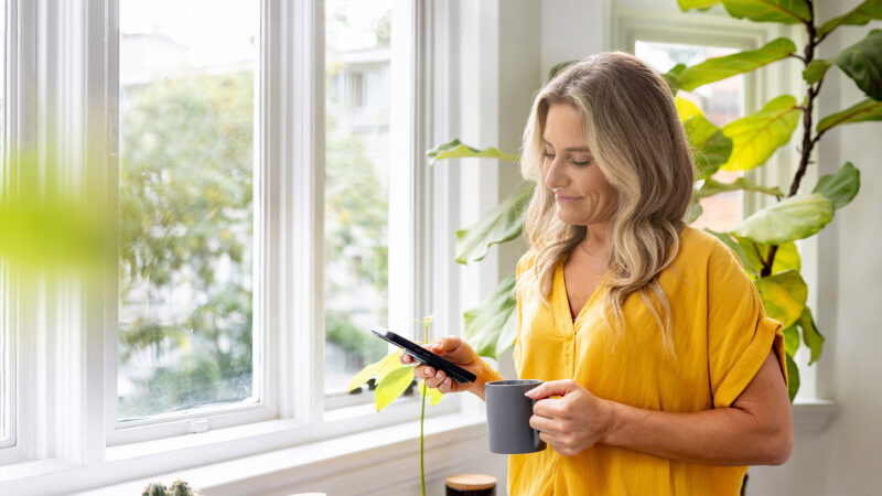 Une femme blonde en haut jaune consulte son smartphone tout en tenant une tasse grise près d'une fenêtre lumineuse.