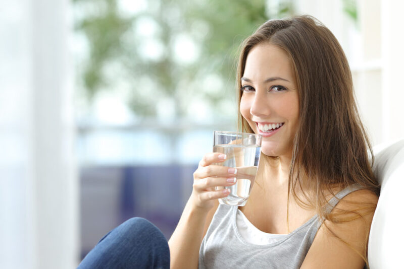 Une femme souriante tient un verre d'eau transparente près de ses lèvres.